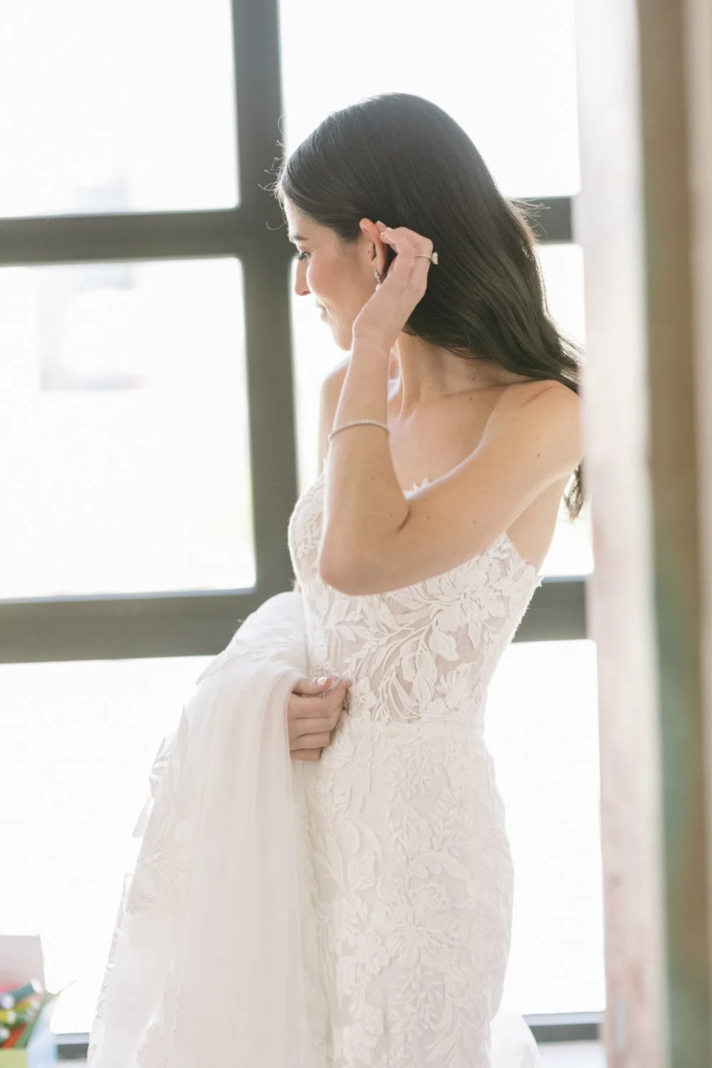 A bride in a white lace wedding dress adjusting her hair near a large window.