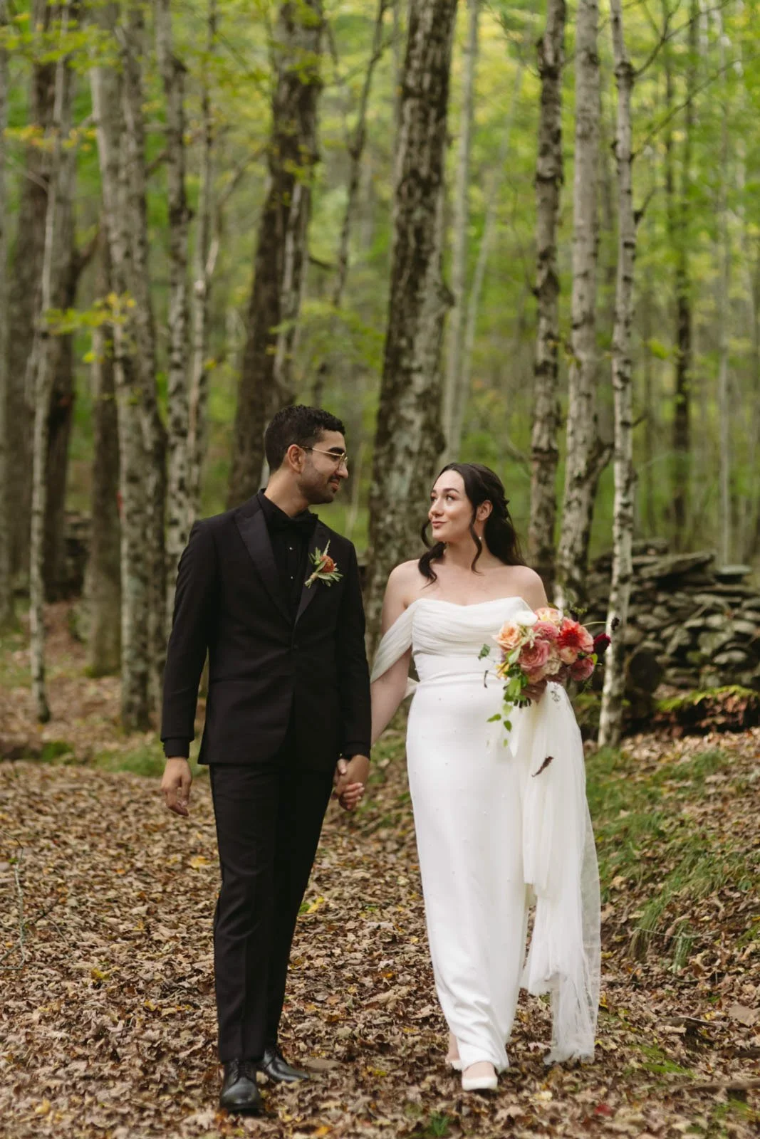 A bride and groom walking hand in hand through a forest with green trees, the bride holding a bouquet of pink and red flowers.
