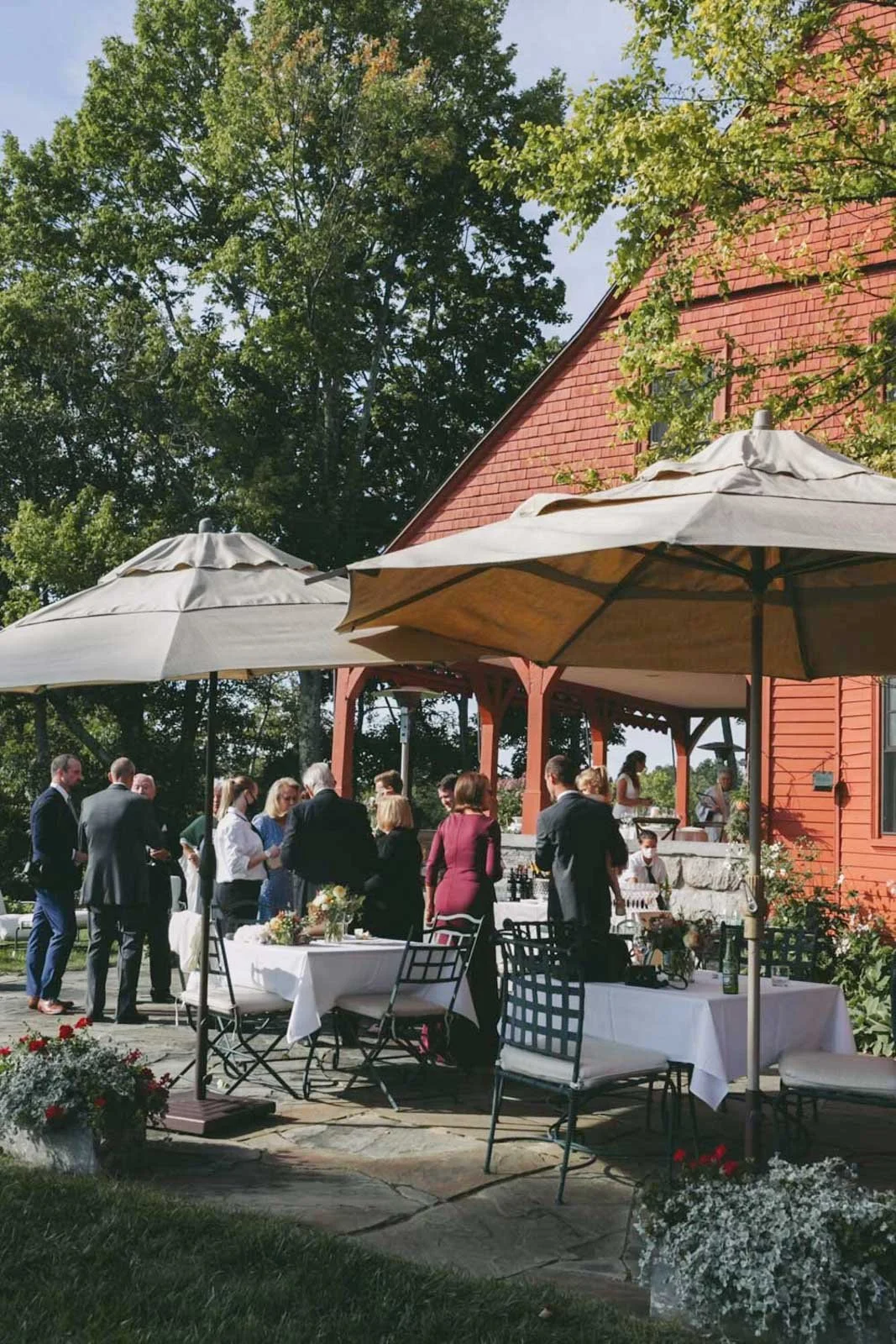 Gathering of people outdoors on a sunny day with tables, chairs, umbrellas, and a red barn-style building in the background.