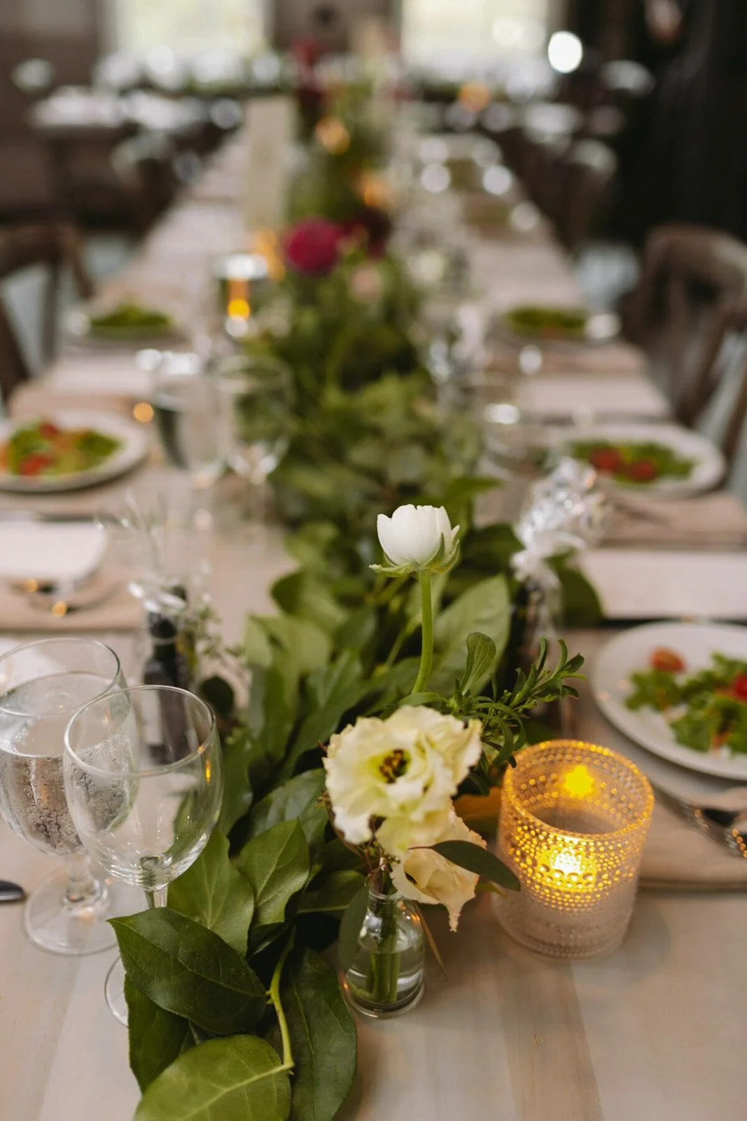 A long dining table set for a meal with a floral centerpiece, wine glasses, plates of salad, and a lit candle, in a well-lit room.