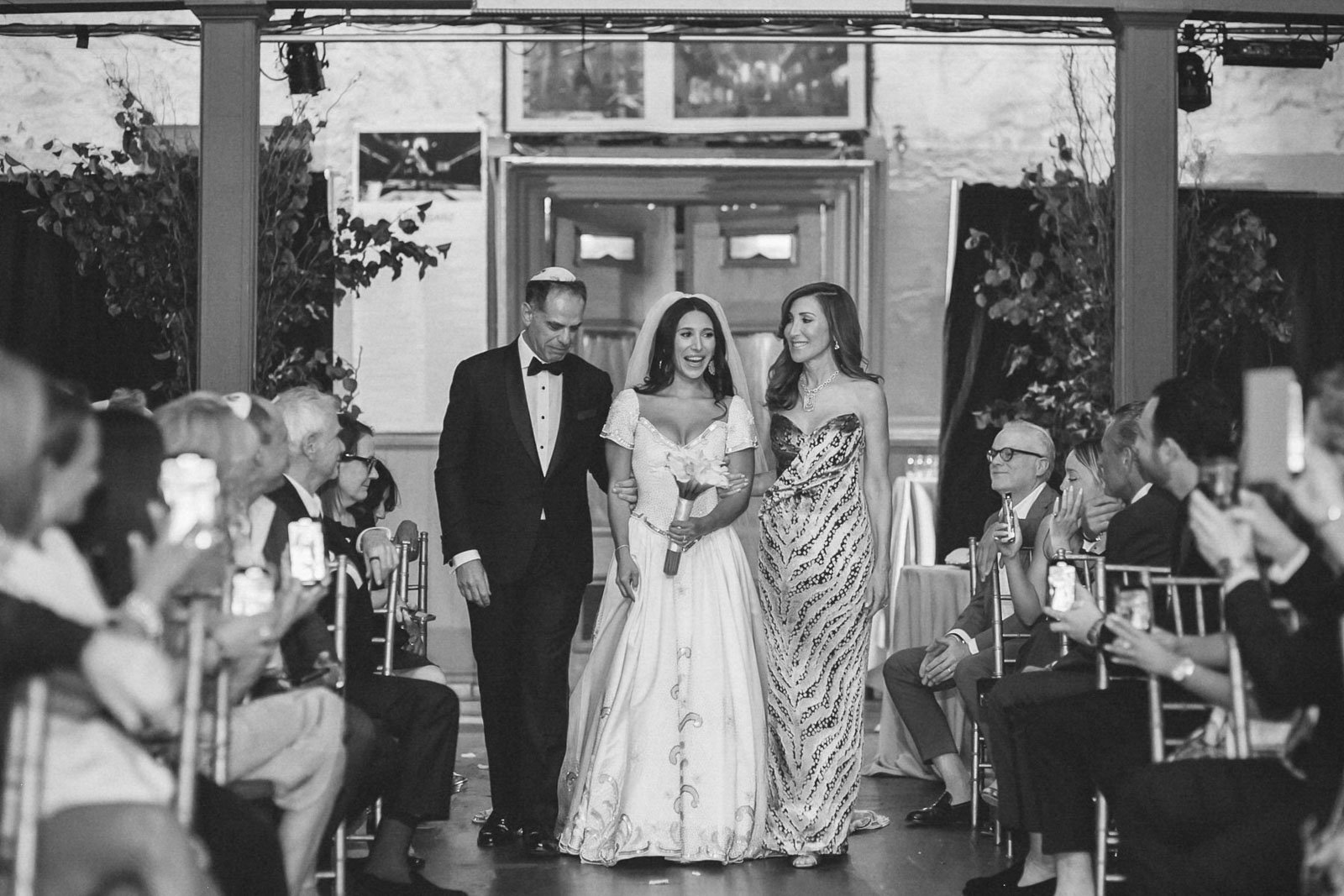 A bride walking down the aisle during her wedding, accompanied by her father and a woman, with guests seated on either side taking photos in a decorated indoor venue.