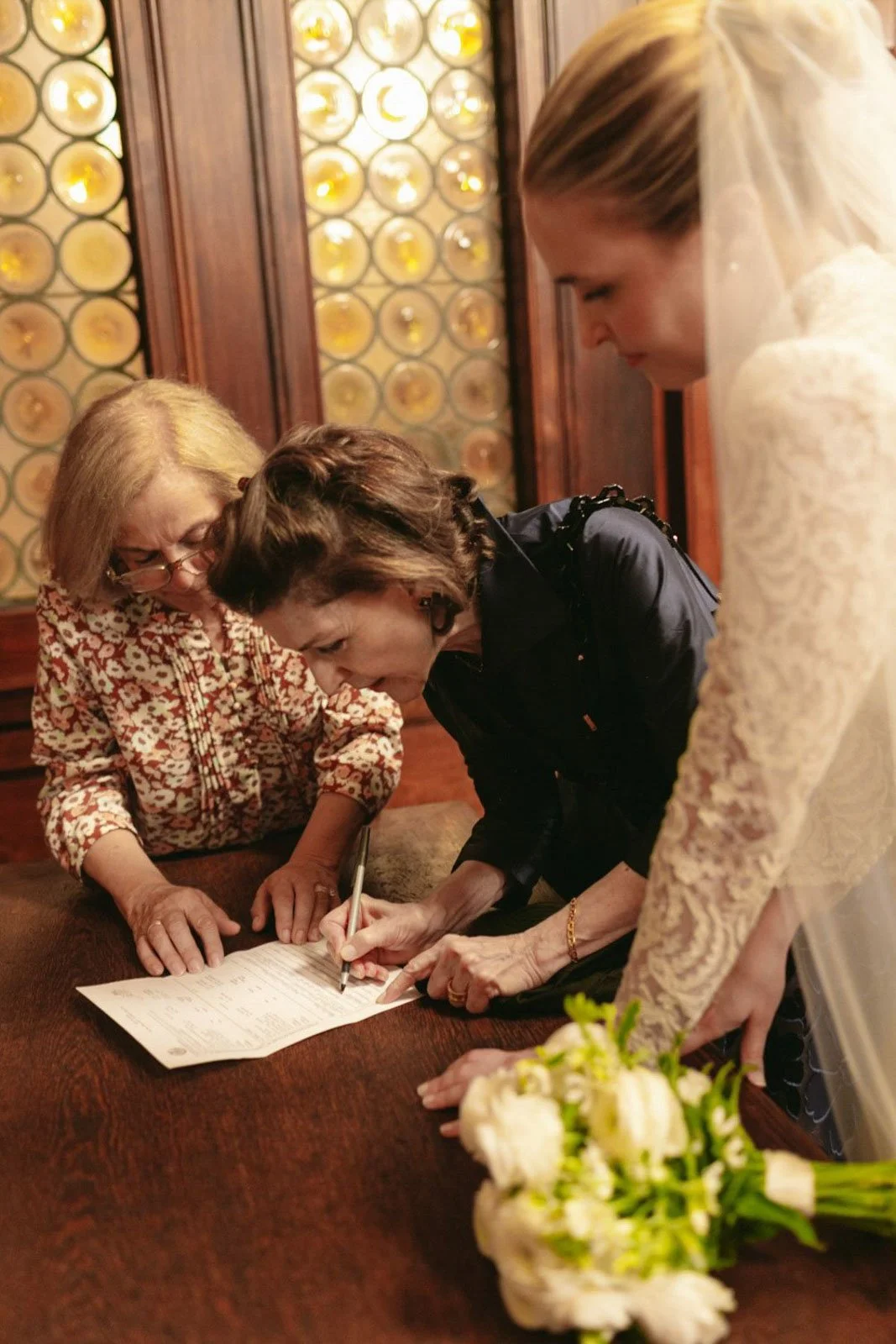Three women signing a document at a table, with a bouquet of flowers nearby.
