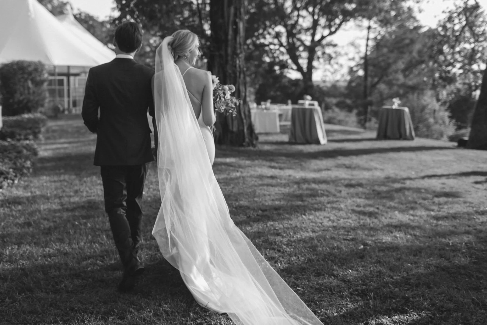 A bride holding a bouquet and wearing a veil walking outdoors with a groom dressed in a suit, surrounded by trees, with tables in the background, in black and white.