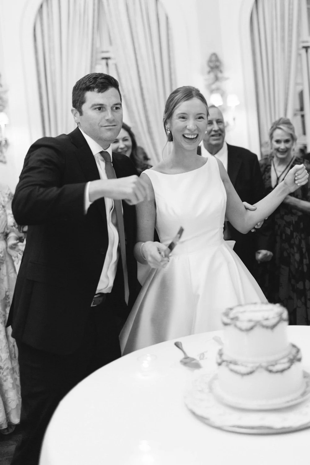 A black and white photo of a wedding celebration showing the bride and groom cutting the wedding cake together, surrounded by smiling guests.
