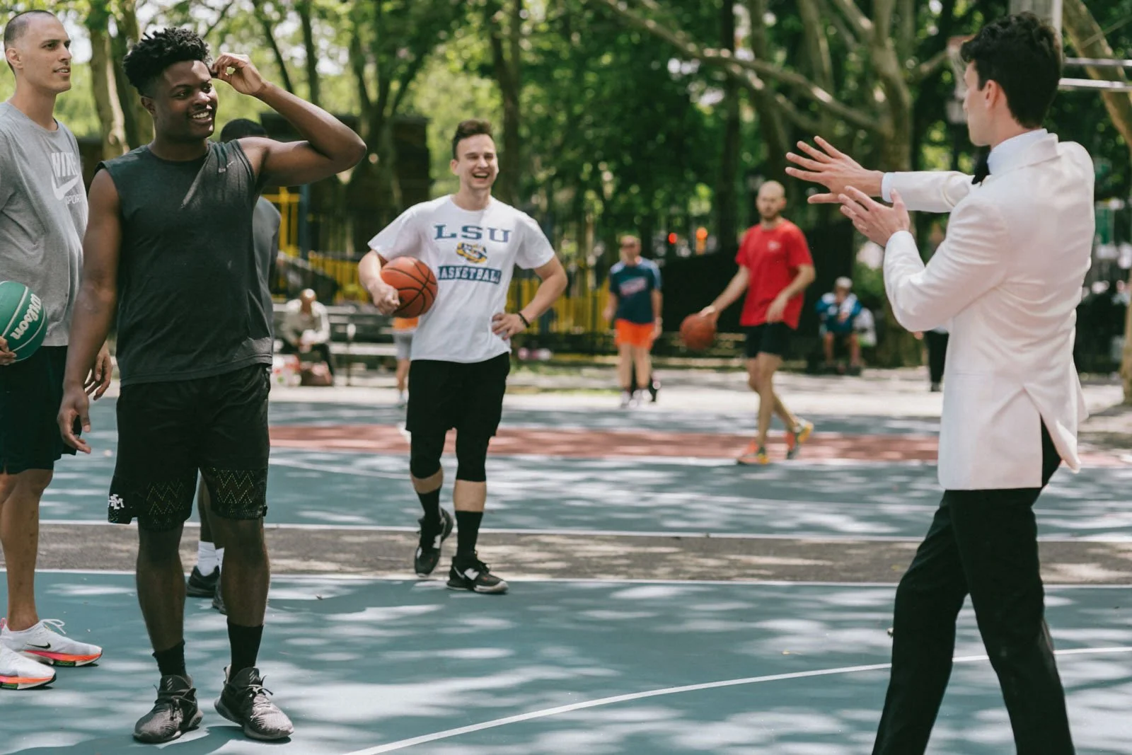 A group of young men playing basketball on an outdoor court, with a man in a white tuxedo shirt coaching or instructing them.
