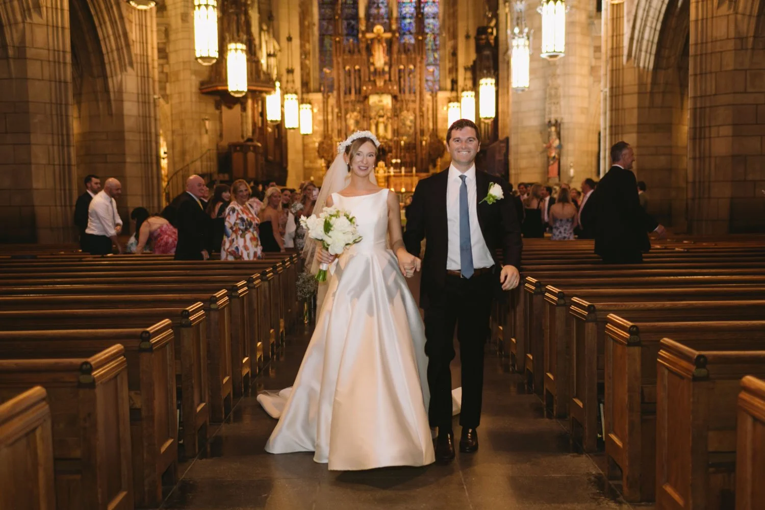 A bride and groom walking down the aisle in a church after their wedding ceremony, smiling and holding hands. The bride is in a white wedding gown holding a bouquet, and the groom is in a dark suit with a boutonniere. Guests are standing and clapping
