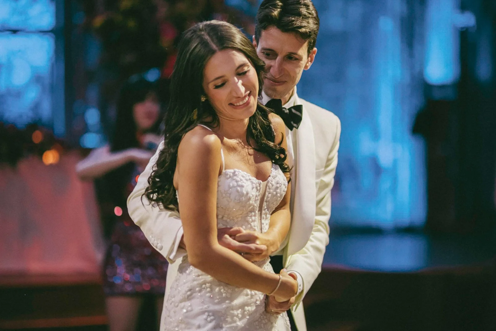 A bride and groom dancing at their wedding reception, smiling and embracing each other in a dimly lit venue.