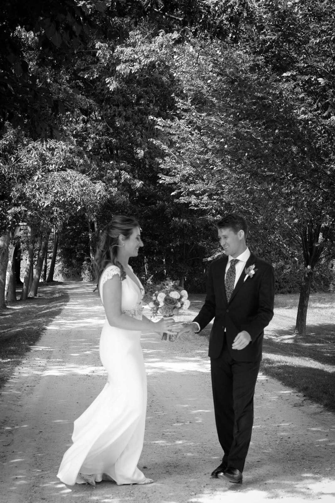 A bride and groom exchanging rings outdoors on a tree-lined path in black and white.