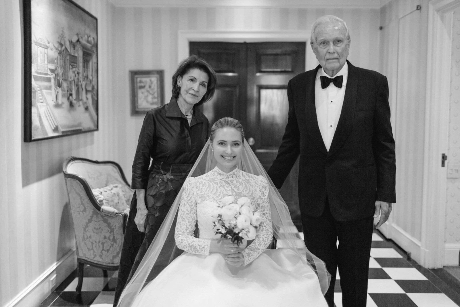 A black and white photo of a bride sitting with her family indoors. The bride is smiling, holding a bouquet, and wearing a lace wedding dress with a veil. Two women and two men are standing behind her, dressed formally.