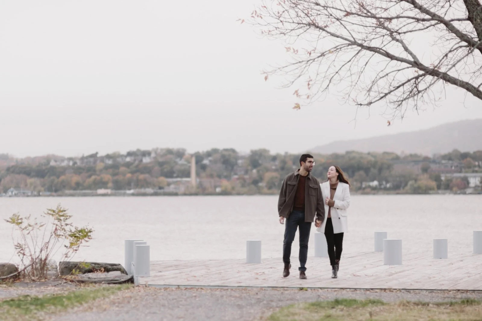 A couple walks hand in hand along Long Dock Beacon, with a river and hilly landscape in the background during autumn, overcast sky, and a tree overhead.