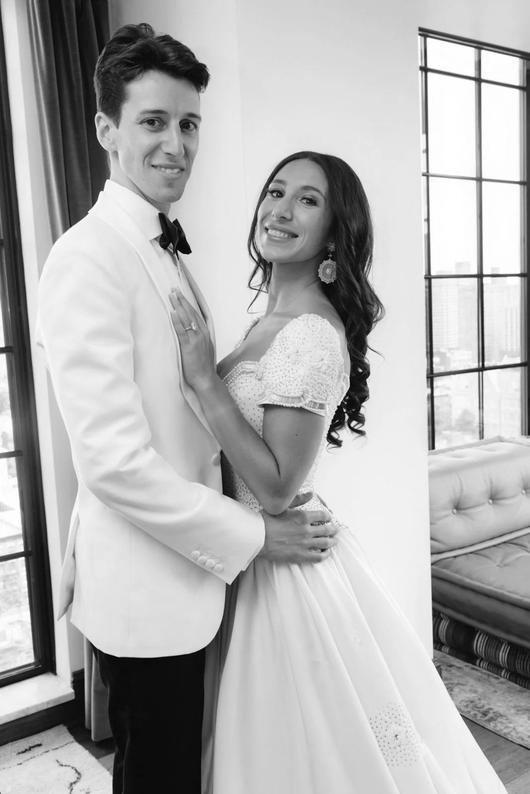 Black and white photo of a bride and groom in wedding attire, standing indoors near large windows with city buildings outside, smiling and holding each other.