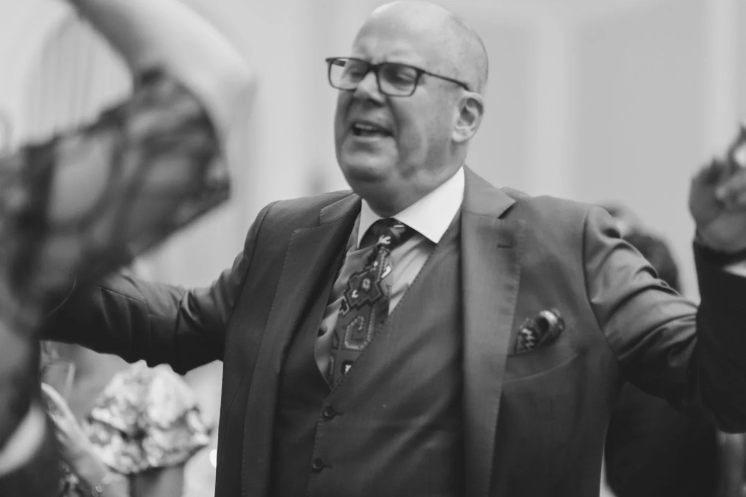 A man wearing a suit, glasses, and a tie, passionately speaking or singing with his arms raised in a lively indoor setting.