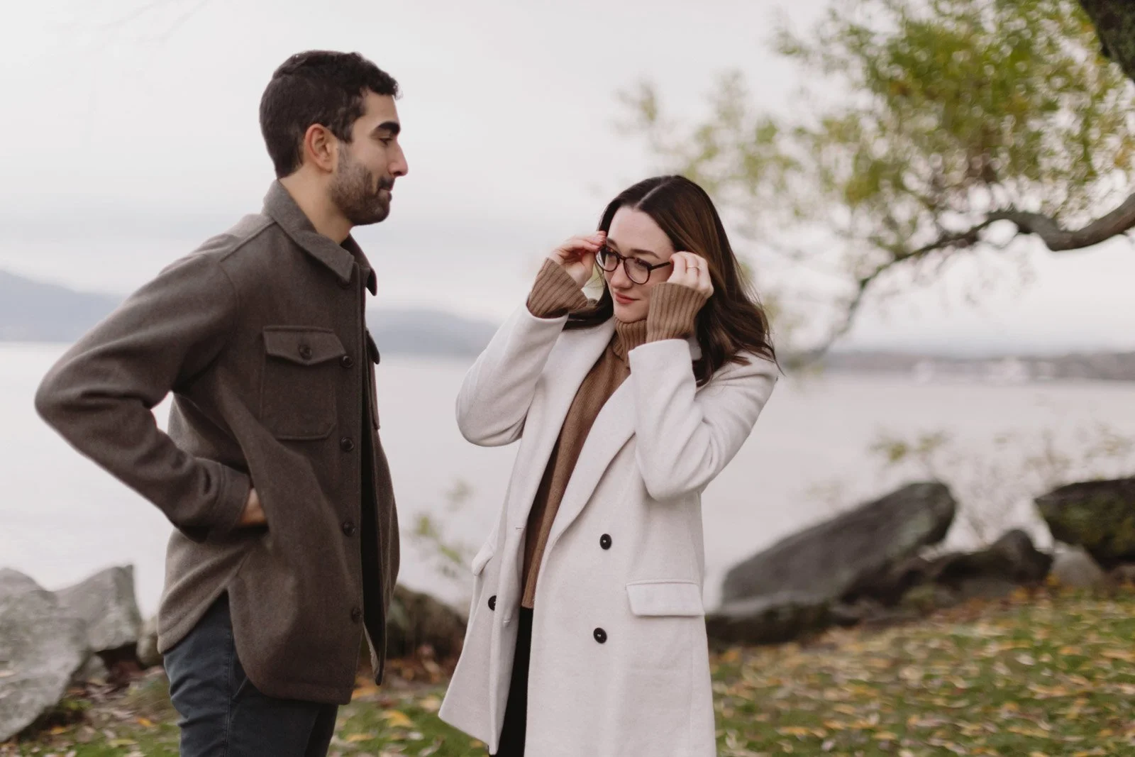 A man and a woman standing outdoors near water, engaged in a conversation. The woman is adjusting her glasses, and the man is looking at her. They both are wearing coats, and there are trees and rocks in the background.