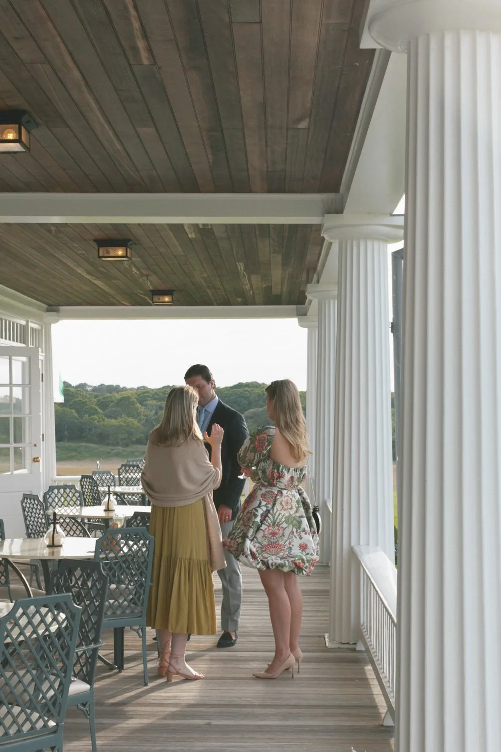 Three people gather on a porch overlooking a green landscape, engaged in conversation. The porch has wooden floors, white columns, and tables with black lattice chairs, with a partially open door on the left.