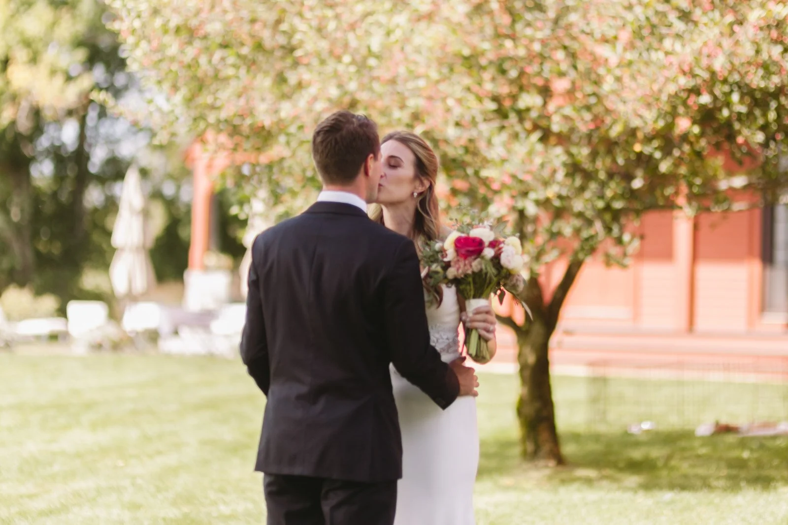 A bride and groom kissing outdoors on their wedding day, with the bride holding a bouquet of flowers and a tree with pink blooms in the background.