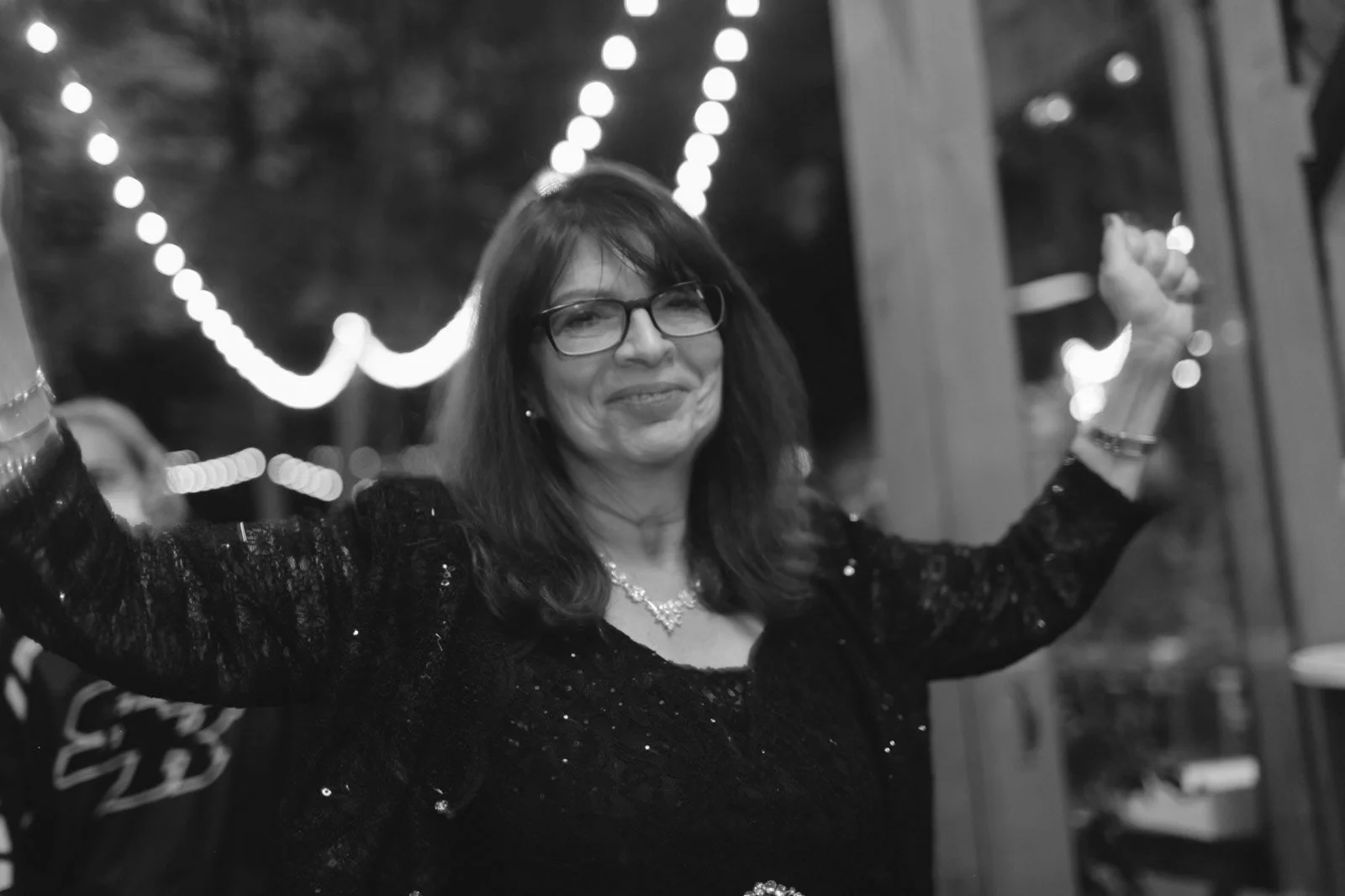 A woman with glasses and dark hair, wearing a black sequined dress and jewelry, celebrating at a party with her arms raised, with string lights in the background.