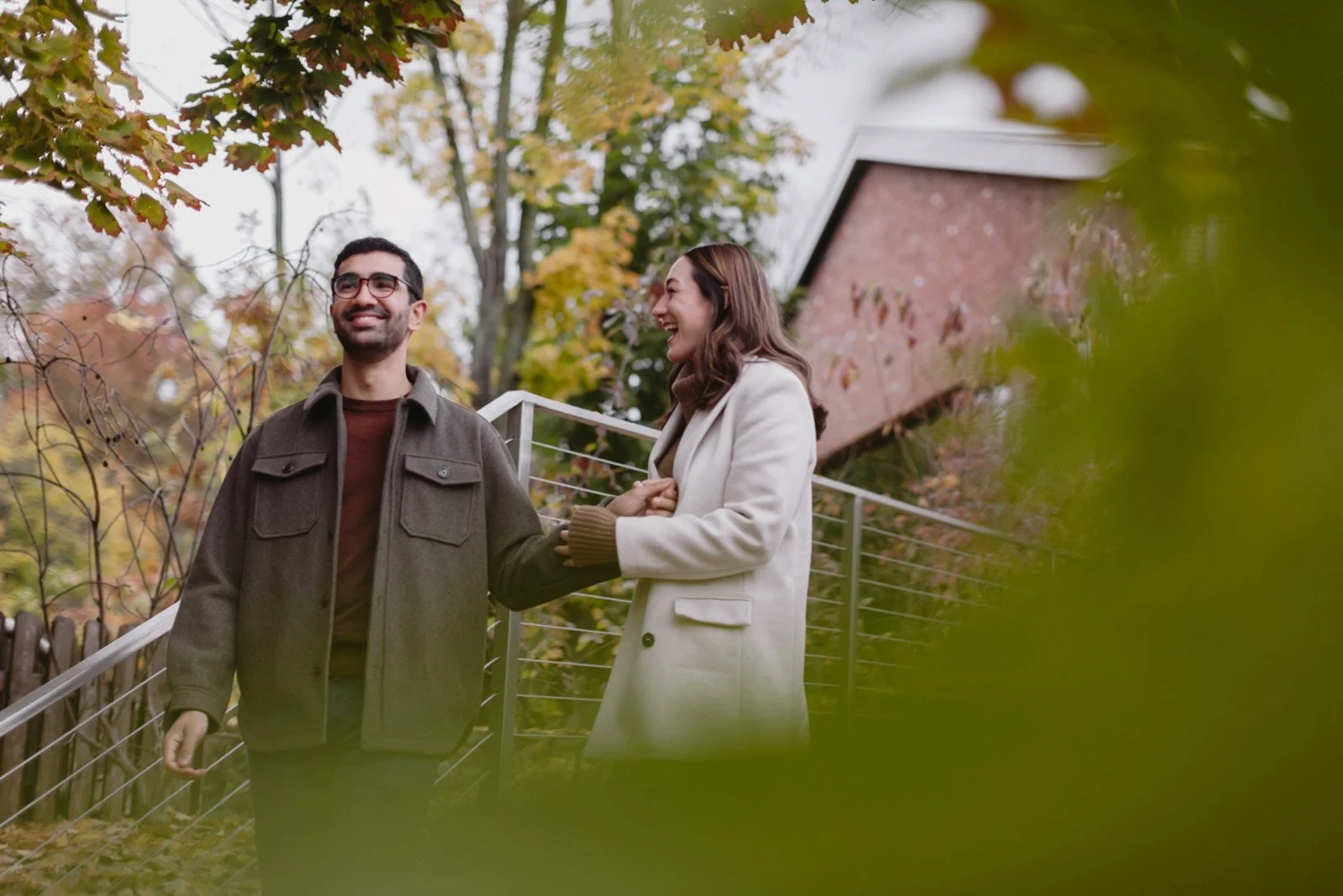 A smiling man and woman walk outdoors on a fall day, holding hands, surrounded by autumn leaves and trees with colorful foliage.
