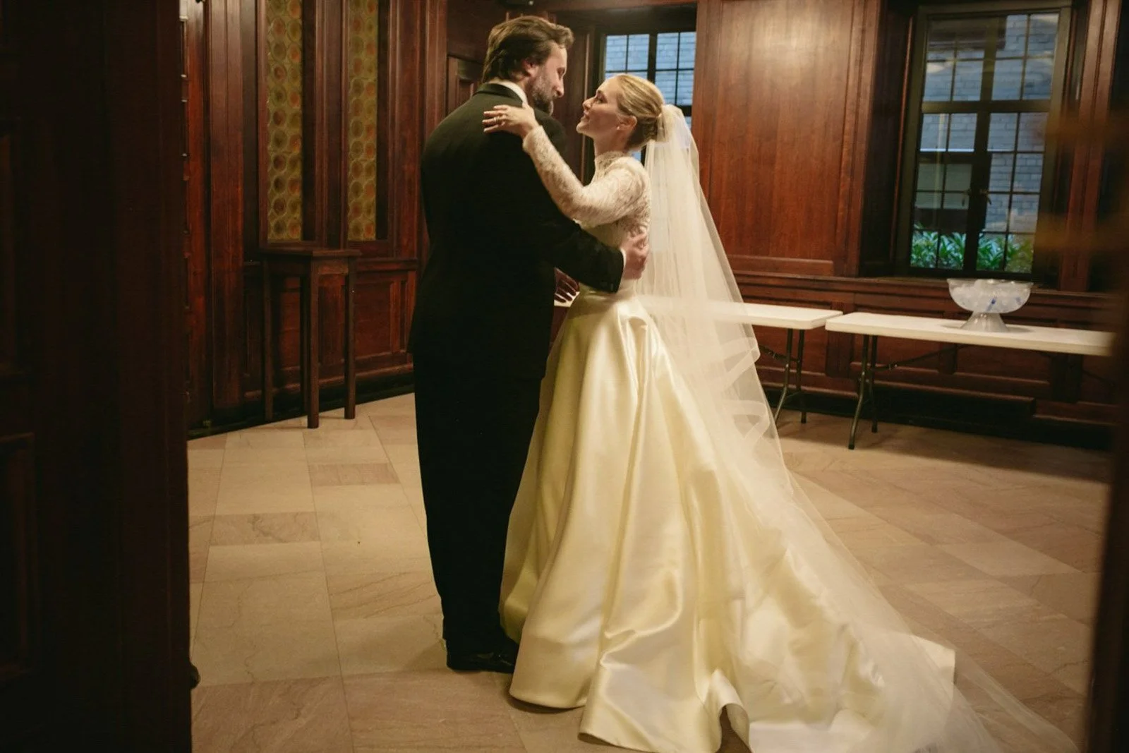 A bride and groom dancing together in a warmly lit wood-paneled room, with the bride wearing a long white satin wedding gown and veil, and the groom dressed in a dark suit.