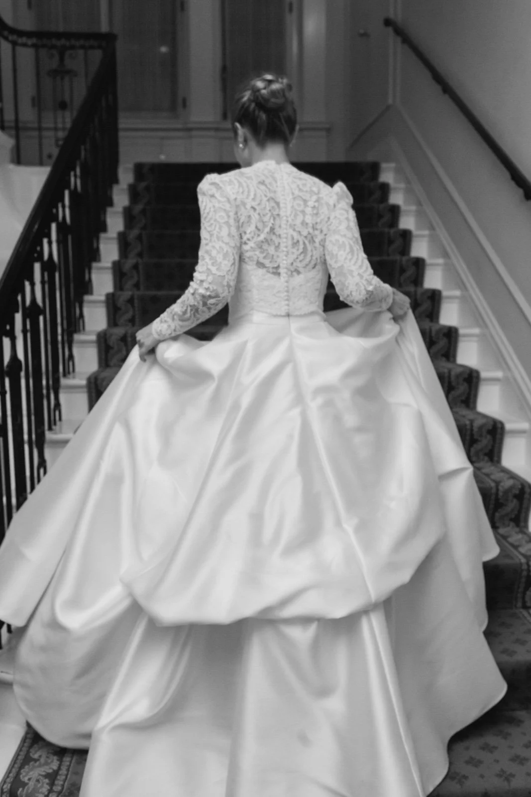 A woman in a wedding gown ascending a staircase, holding the sides of her long satin train with lace detailing on the upper part of the dress at The Colony Club New York.