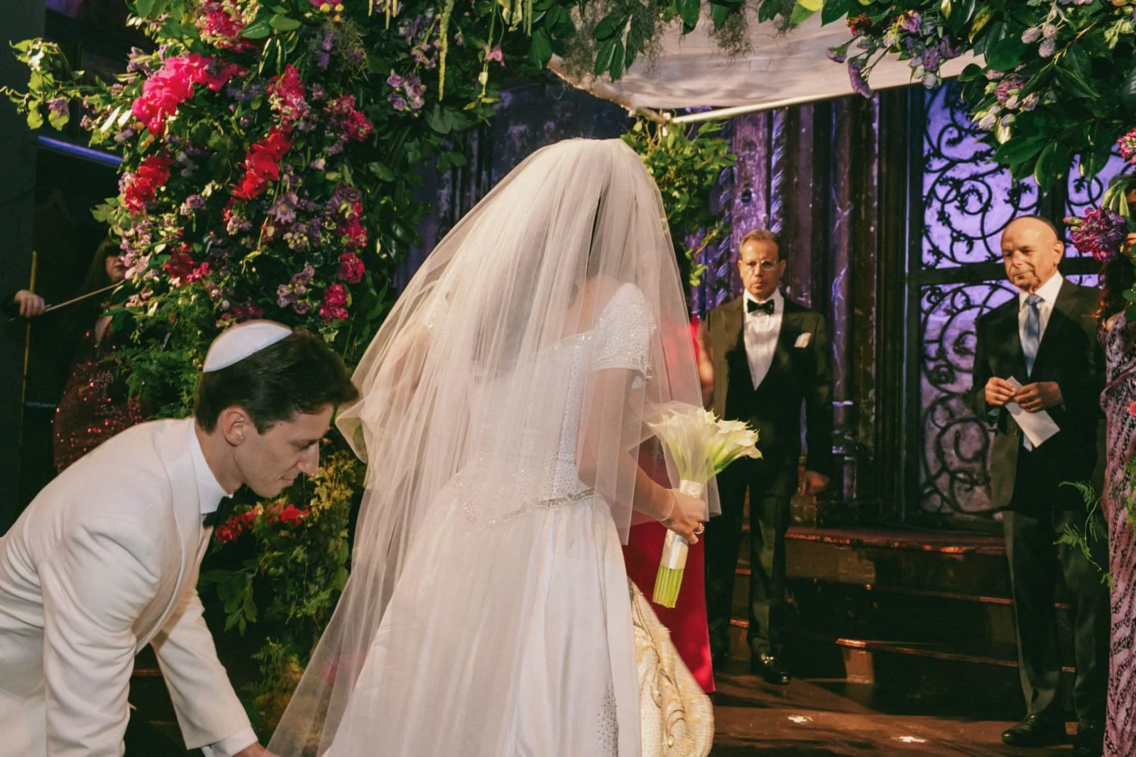 A bride in a wedding dress and veil holding a bouquet of white calla lilies standing next to a groom in a white tuxedo, surrounded by floral decorations and officiants at a wedding ceremony.