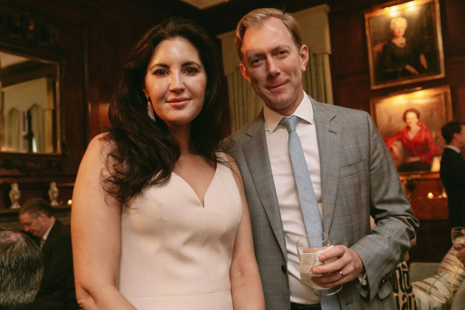 A woman in a white dress and a man in a gray suit pose together at a social gathering in a warmly lit room with wooden paneling and framed portraits on the wall.
