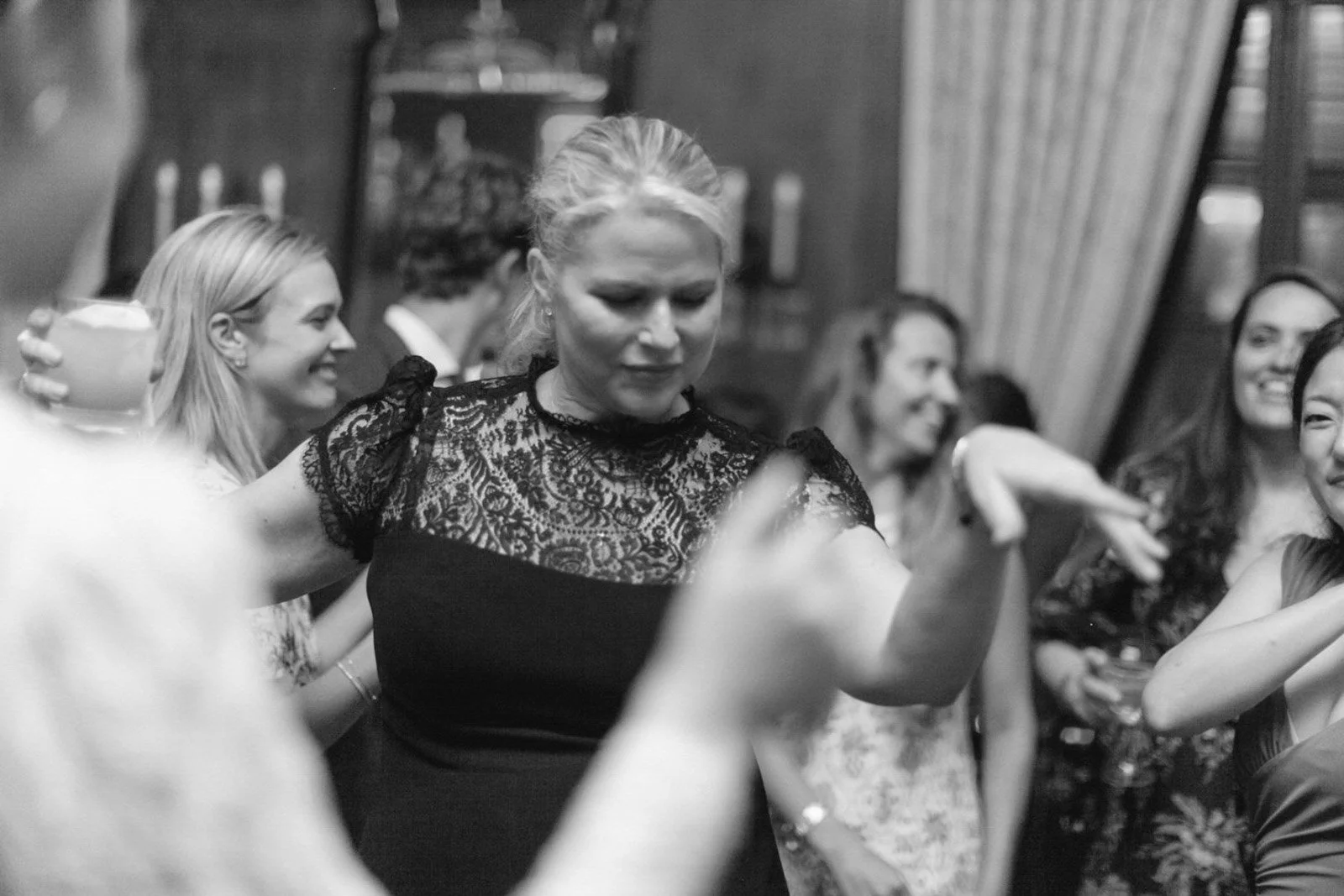 A group of women dancing and smiling at a social gathering or party, with one woman in the center wearing a black lace dress.
