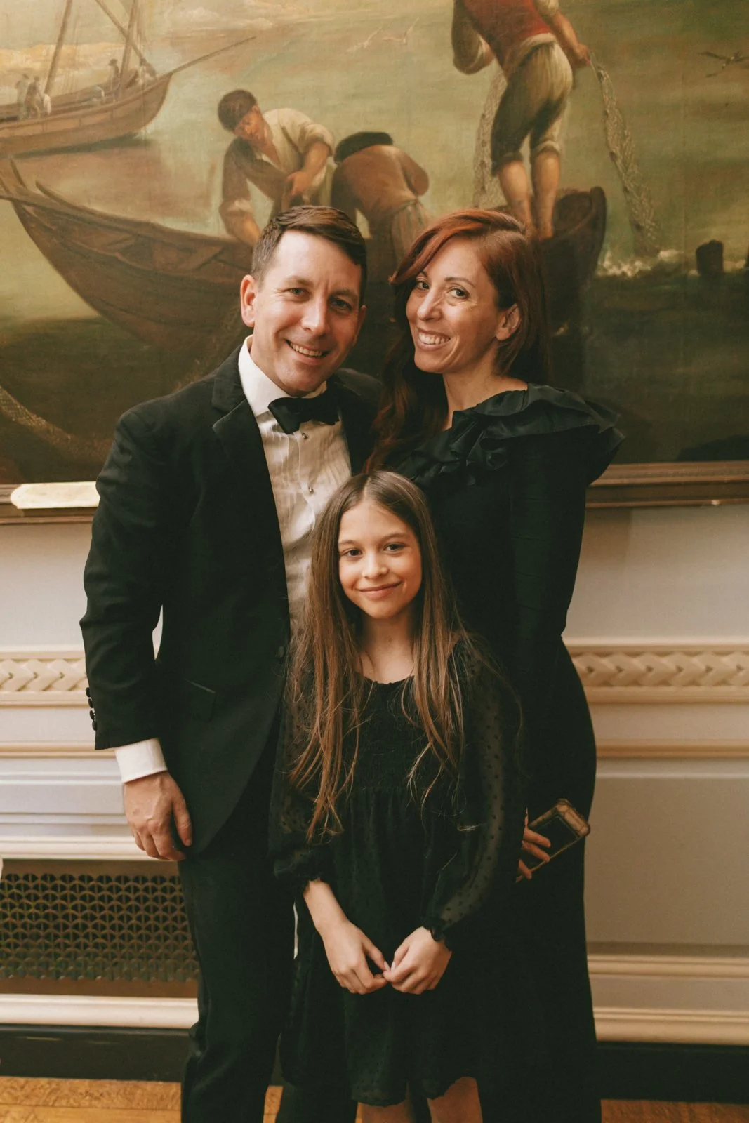 A family of three standing in front of a large historical painting. The man is wearing a tuxedo and the woman is in a black dress, while the girl is dressed in black with long brown hair.