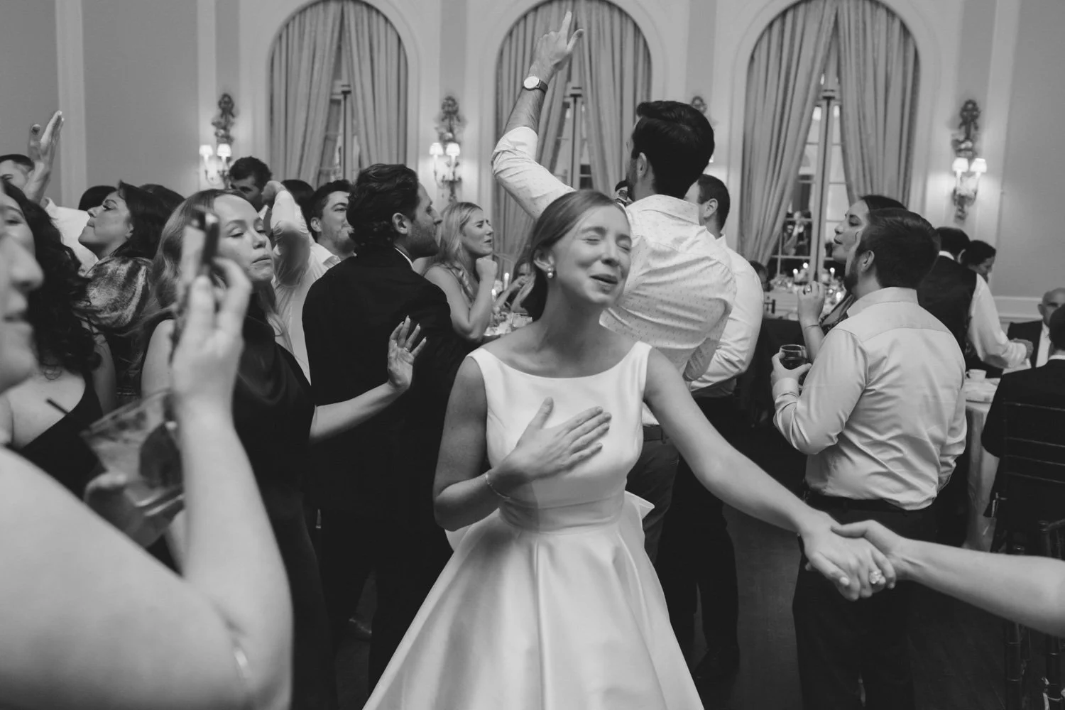 A woman in a white dress cries with her hand on her chest, holding hands with someone at a dance at a formal event in an elegant room filled with other dancing couples.