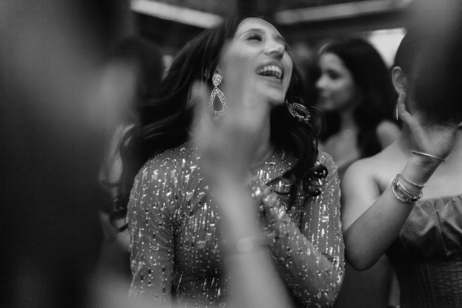 A woman with long dark hair and large earrings is smiling and laughing, wearing a embellished dress with sequins, in a social gathering.