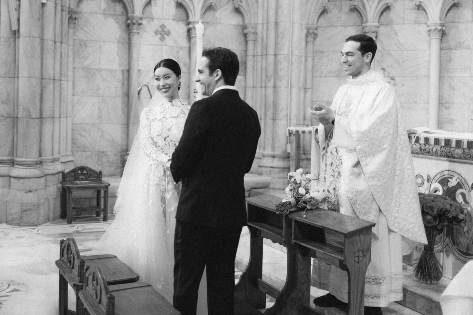 A black and white photo of a wedding ceremony inside a church, with a bride in a lace wedding gown and a veil, a groom in a dark suit, and a priest in ceremonial robes officiating the wedding.