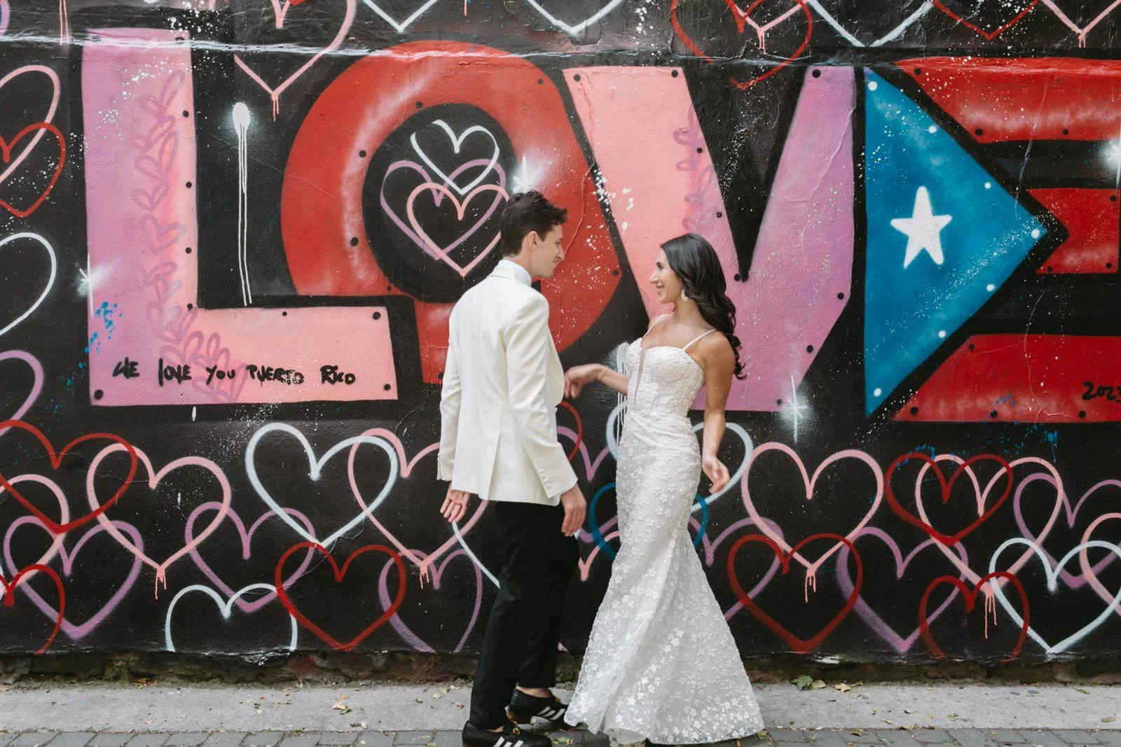 A couple in wedding attire holding hands and looking at each other in front of a colorful graffiti wall with hearts and love-themed designs.