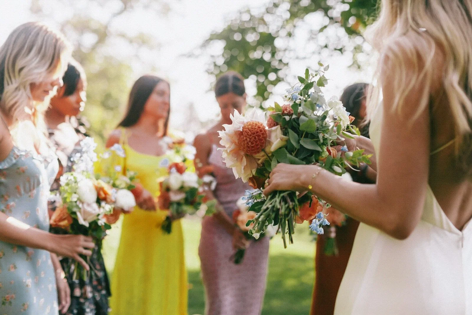 A group of women holding bouquets of flowers at an outdoor event, likely a wedding, with sunlight filtering through trees in the background.