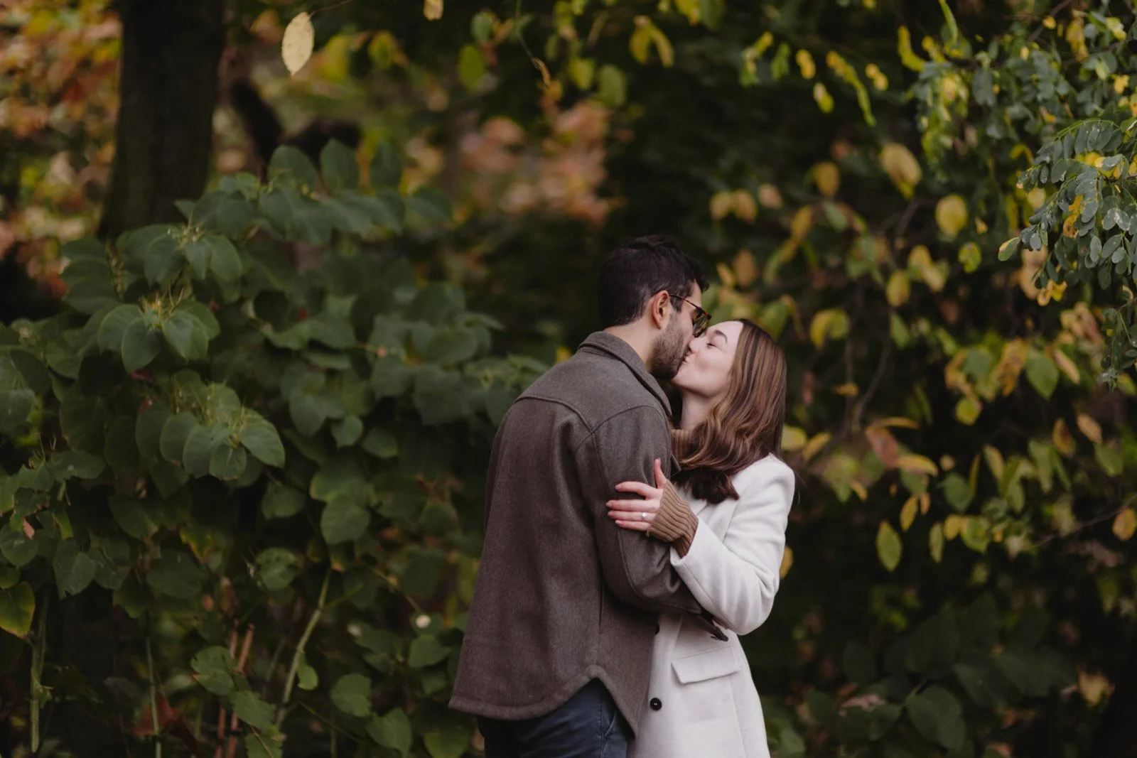 A couple kissing outdoors surrounded by autumn-colored foliage, wearing warm clothing.