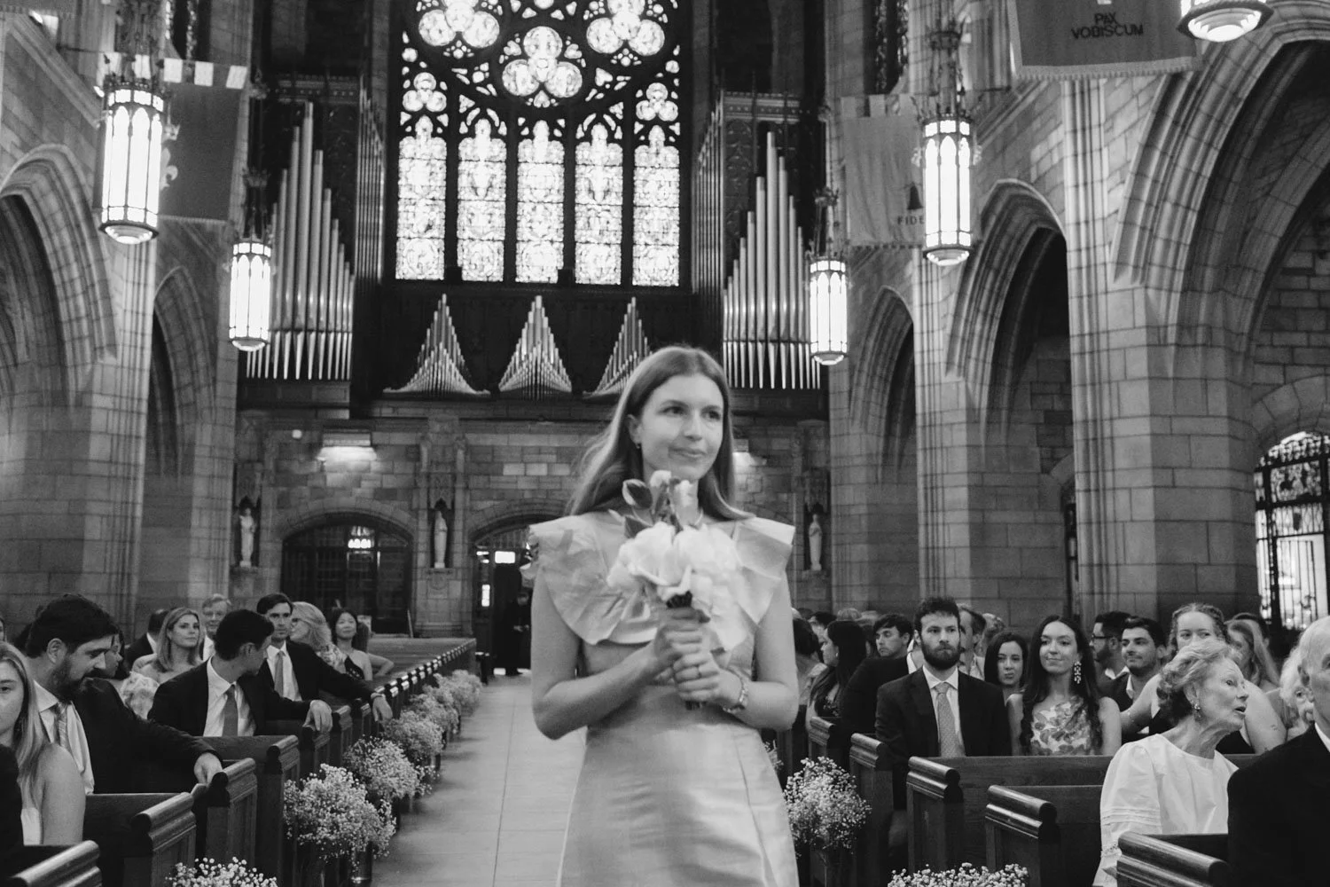 A woman in a wedding dress holding a bouquet, walking down the aisle of a church during a wedding ceremony, with guests seated on benches on either side.