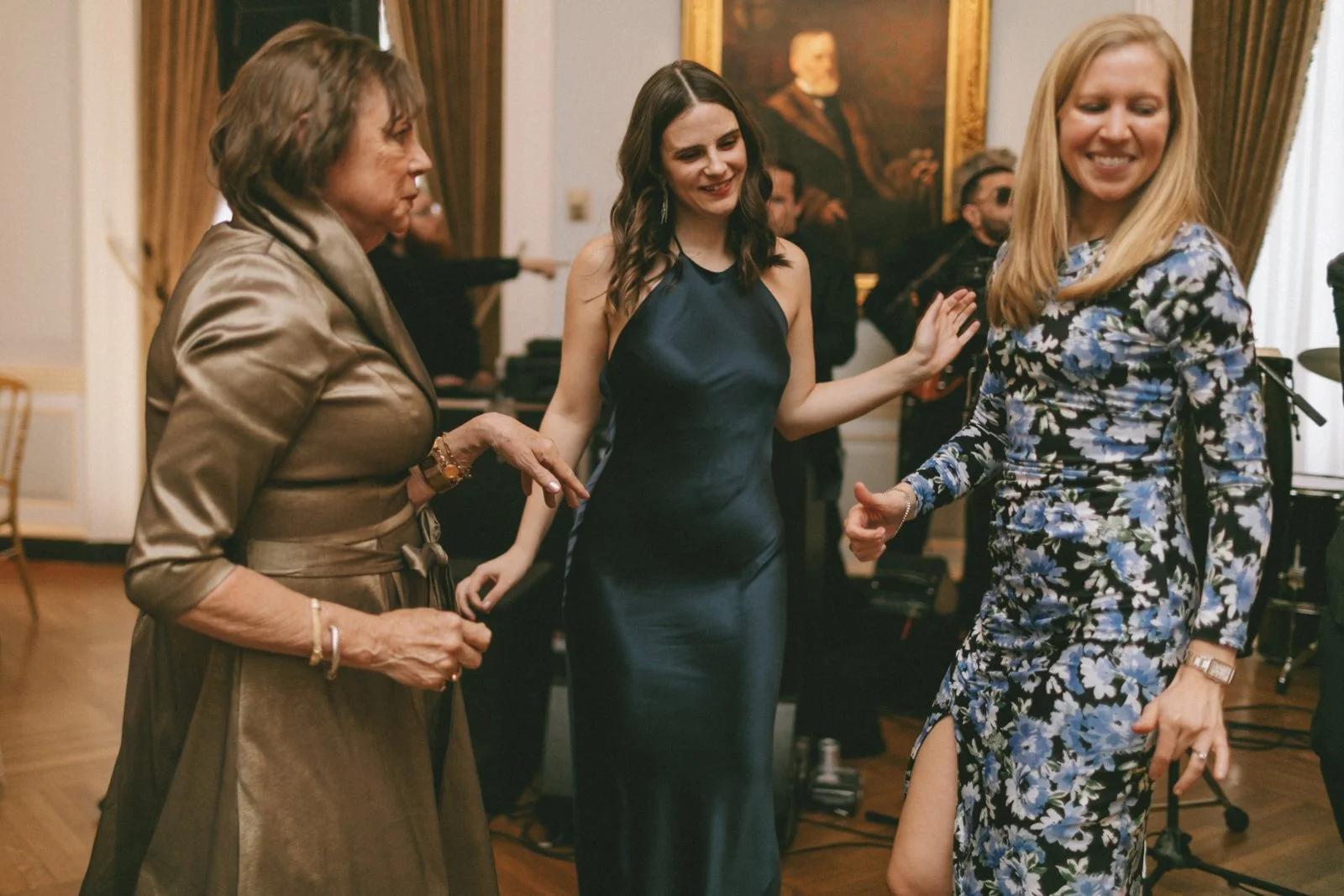 Three women dancing and smiling at a social gathering, with a man in sunglasses and musicians in the background.