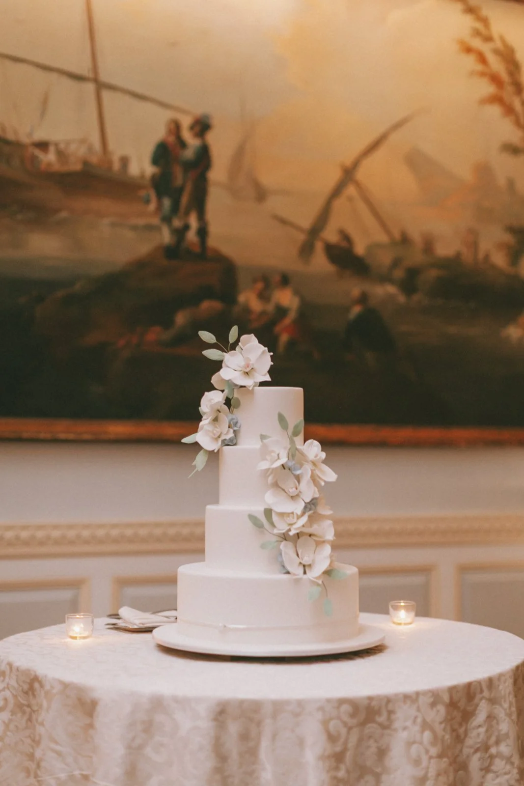 A layered white wedding cake decorated with white flowers and green leaves, placed on a round table with a cloth, with small lit candles on either side. A large framed painting of a historical scene hangs in the background.