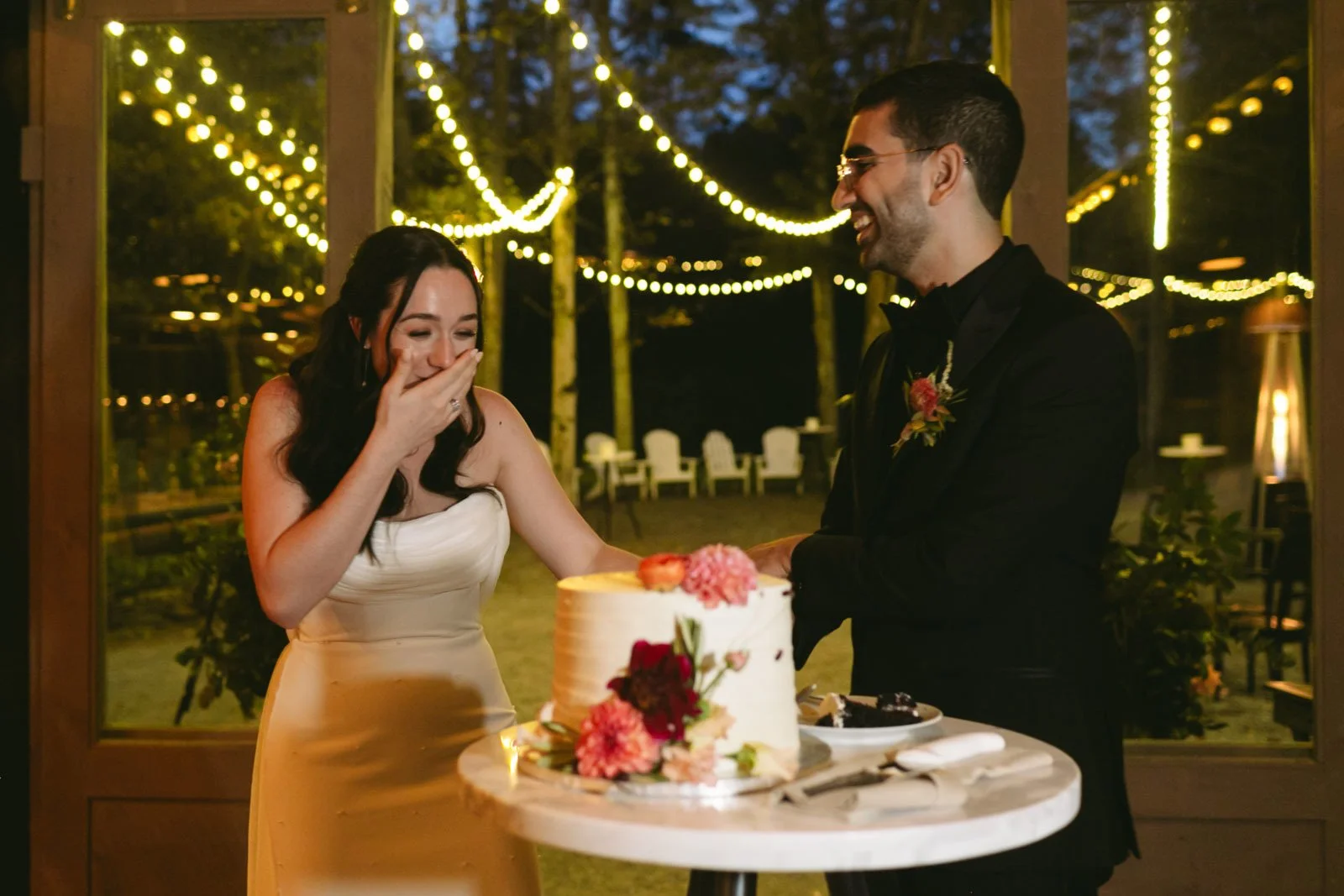 A woman in a white dress and a man in a black tuxedo are at a wedding reception, standing behind a table with a wedding cake decorated with flowers. The woman is covering her mouth in surprise or joy, and both are smiling. String lights are visible o