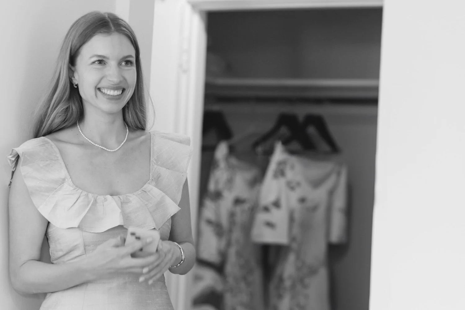 Black and white photo of a woman with long hair smiling and holding a phone, standing in front of an open closet with clothes hanging inside.