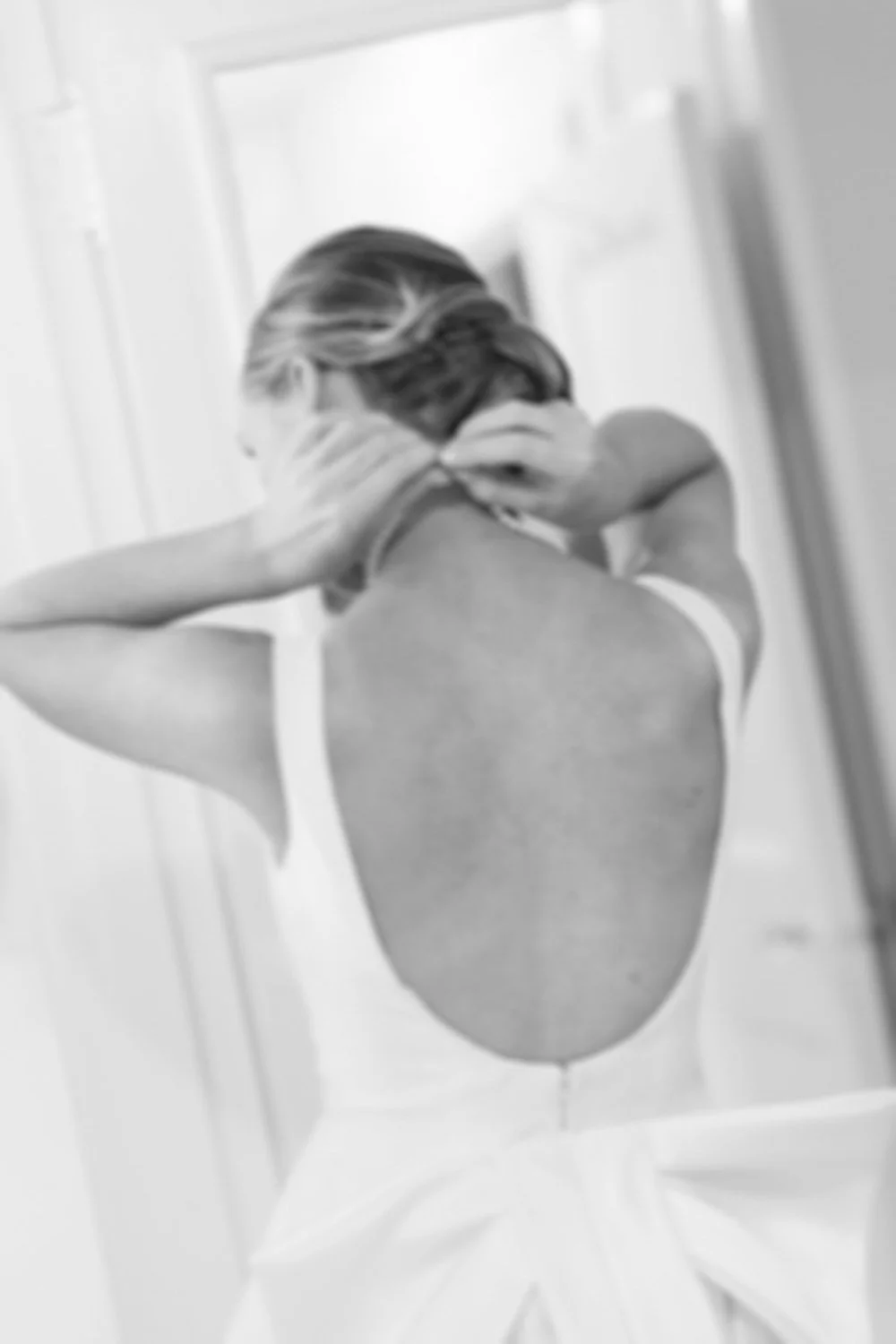 A woman wearing a white dress with a low back, adjusting her hair in front of a mirror.