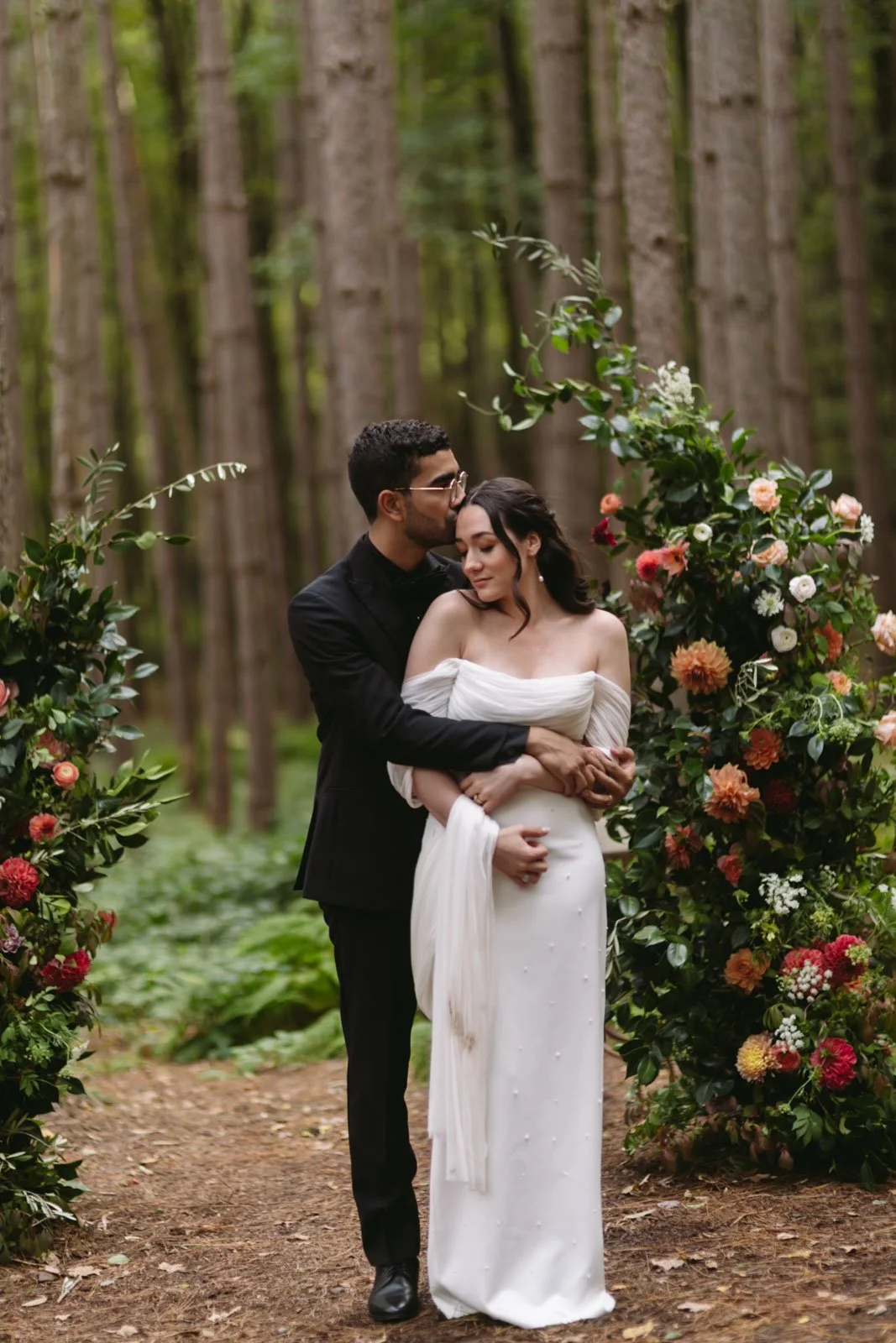 A couple dressed in wedding attire embracing in a forested area, surrounded by floral arrangements at Roxbury Barn and Estate.