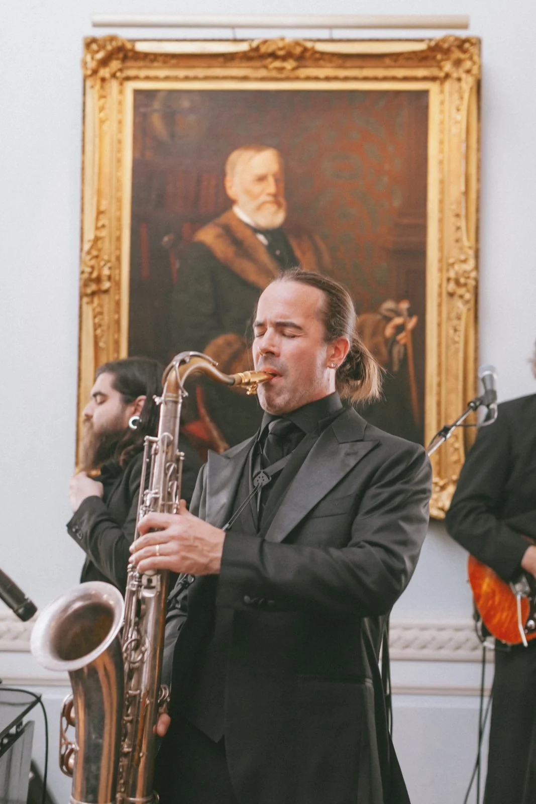 A man in a black suit playing a saxophone in a room with a large framed painting of an older man with a beard in the background.
