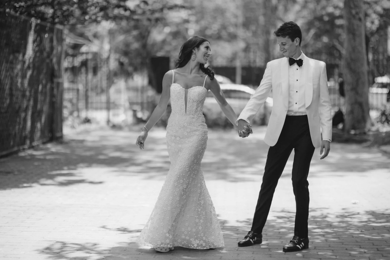 A bride and groom holding hands and walking outdoors, smiling at each other, in a black and white photograph.
