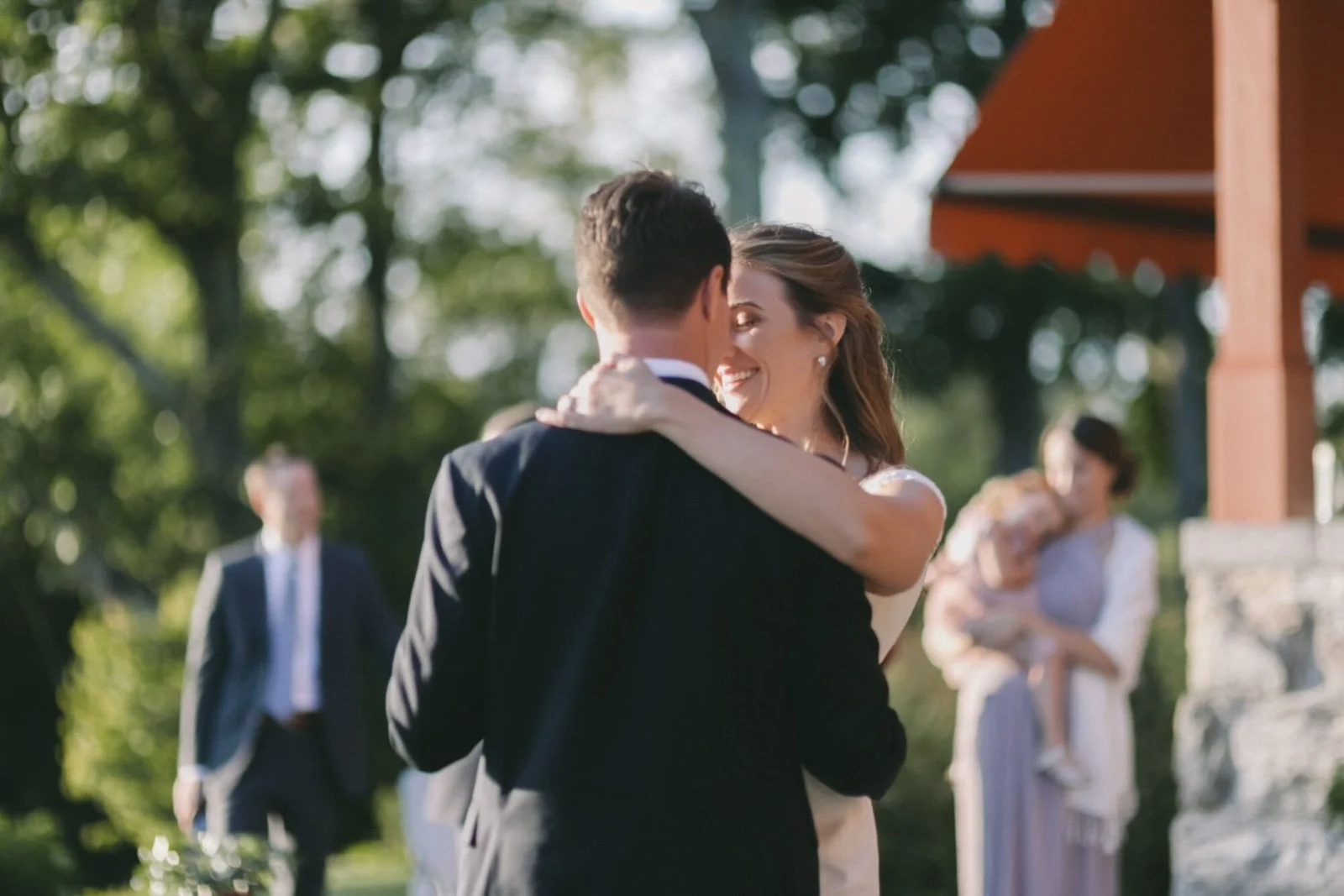 A couple dancing and smiling outside during a wedding or celebration, with a woman holding a child and a man in the background.