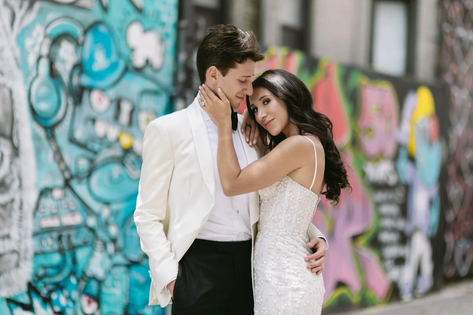 A couple dressed in formal wedding attire, embracing each other in front of a colorful graffiti wall.