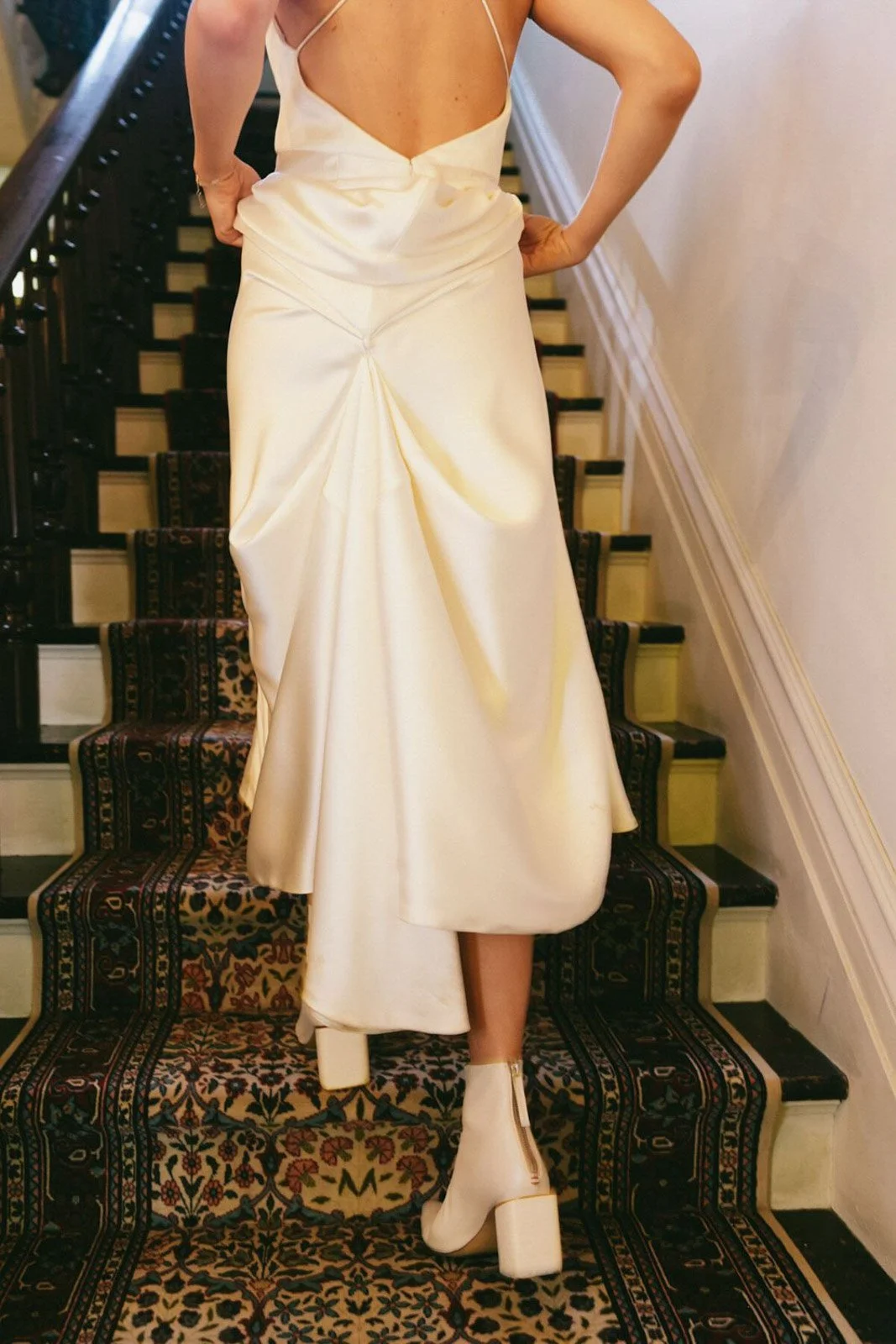 Person in a cream-colored satin dress with a high slit, standing on a staircase with a patterned carpet runner, wearing matching cream boots.