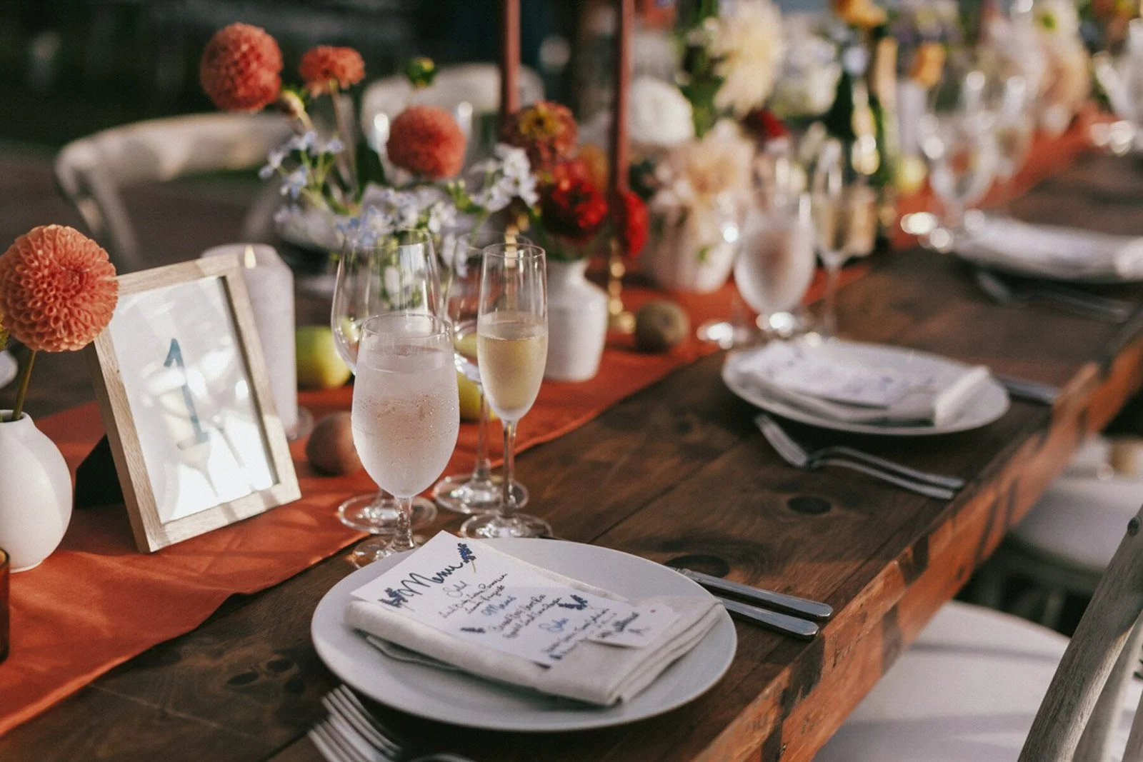A decorated outdoor dinner table with flowers, glasses of water and champagne, place settings with napkins, and a handwritten menu or place card.