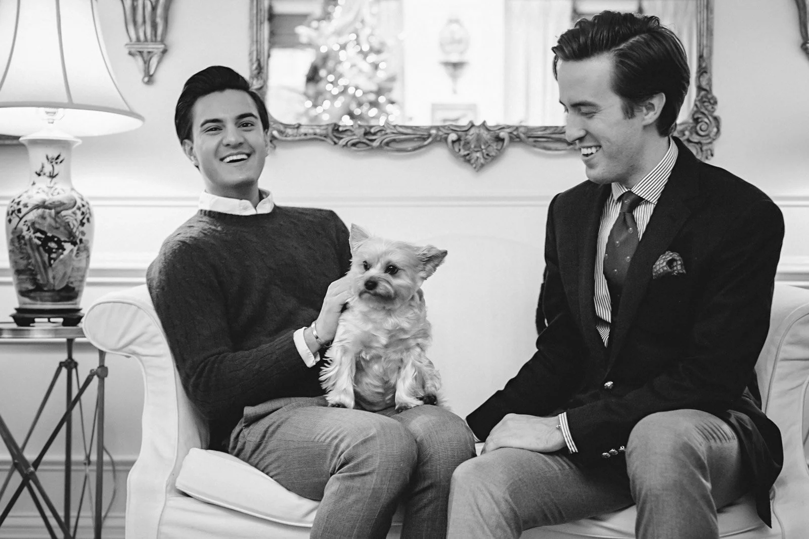 Two young men with dark hair, dressed in formal attire, sitting on a sofa. One is holding a small dog. They are smiling and appear to be in a decorated living room, possibly during a holiday celebration.