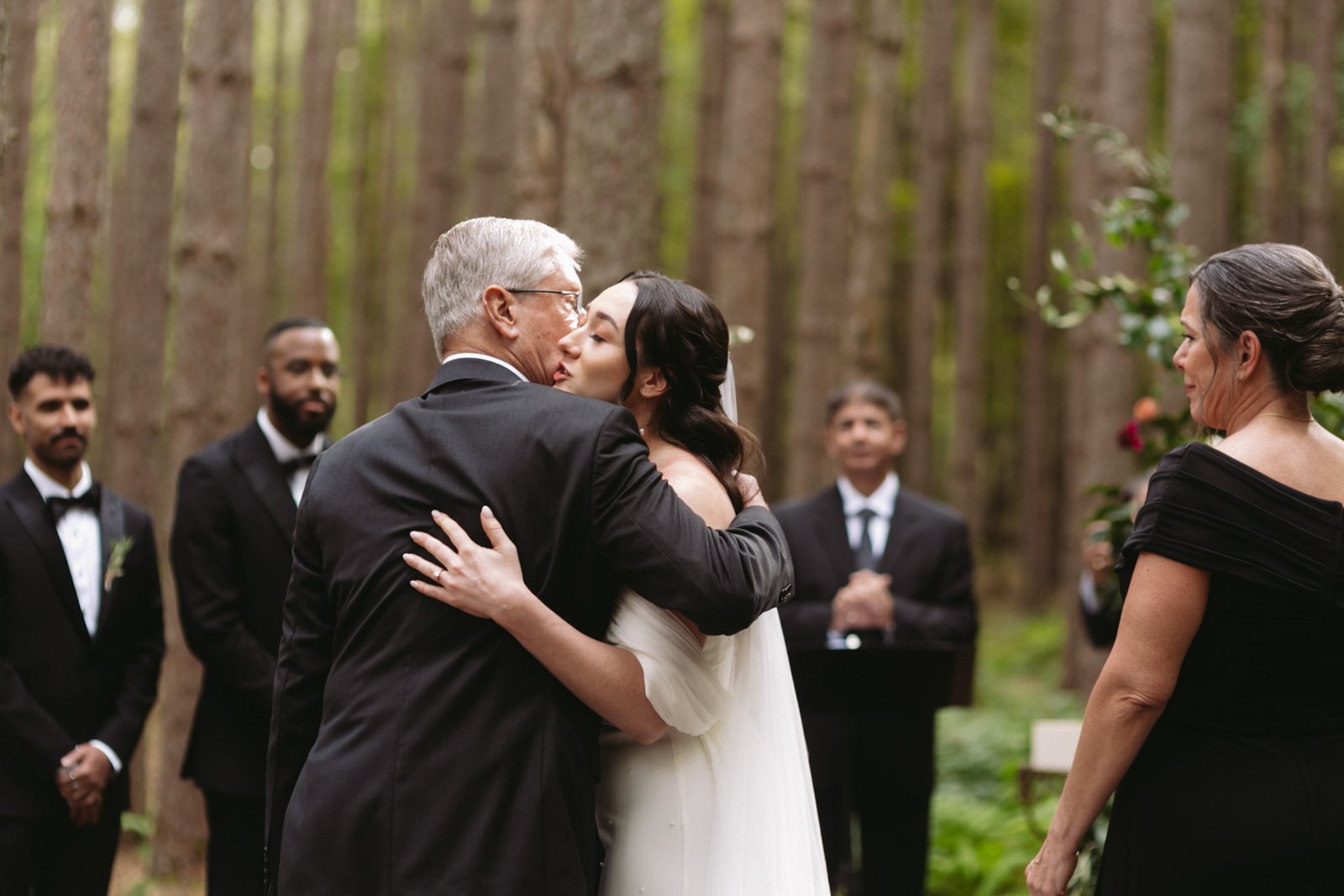 A bride and an older man wearing a tuxedo embrace and kiss during a wedding ceremony in a forest, with two women and three men in tuxedos standing in the background.