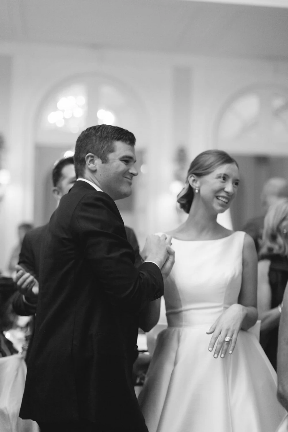 A black and white photo of a wedding dance, showing a man in a black suit and a woman in a white wedding dress smiling and dancing together.
