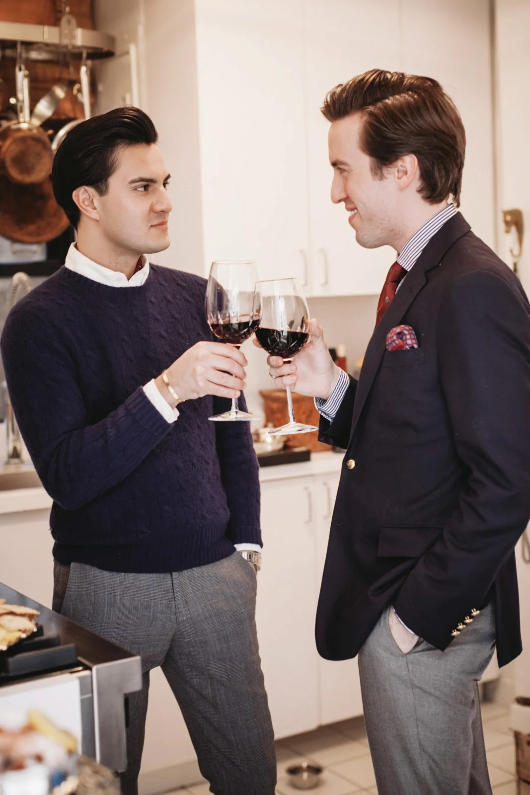 Two men holding glasses of red wine, standing face to face in a kitchen. One is dressed casually in a sweater and plaid pants, the other in a suit with a pocket square. They appear to be engaging in a toast or conversation.